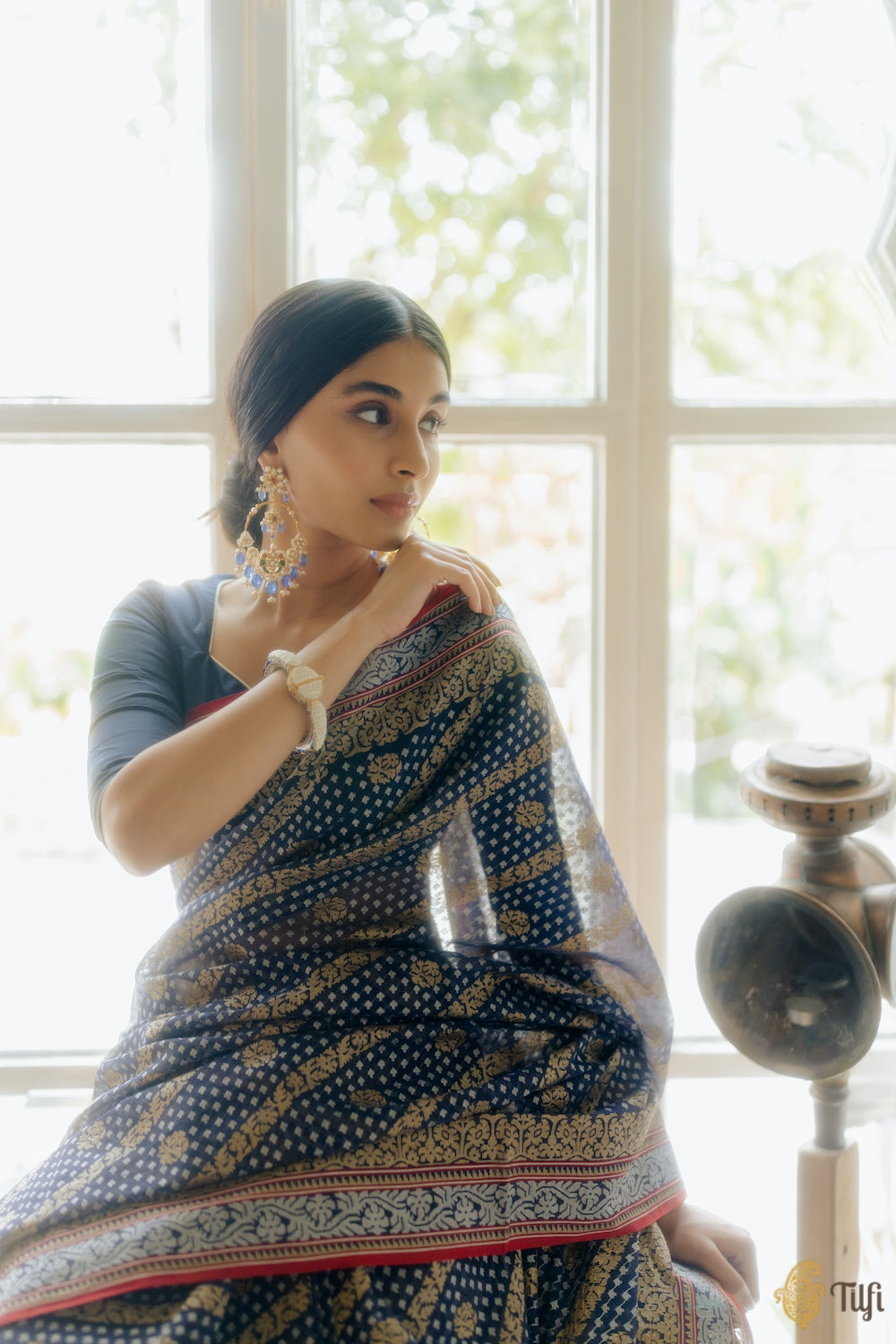 A woman sitting in a room with a navy blue Banarasi Jamdani saree with real zari embellishments and a floral pattern, including corner paisleys, wearing a matching blouse piece.